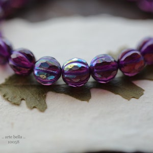 May include: A close-up of a purple iridescent beaded bracelet. The beads are faceted and have a striped pattern. The bracelet is lying on a white surface with a brown leaf.