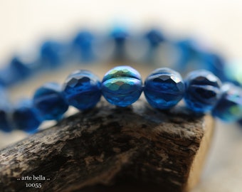 May include: A close-up of a bracelet with faceted, iridescent blue beads. The beads are strung together, creating a circular shape. The bracelet is resting on a piece of wood. The text ".. arte bella .. 10055" is visible.