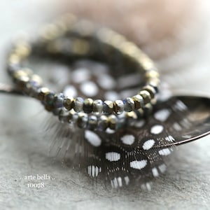 May include: Two strands of beaded jewelry with gold and silver beads on a spotted feather background. The text "... arte bella ... 10078" is visible in the lower left corner of the image.