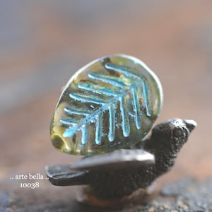 May include: A clear, leaf-shaped bead with a blue fern design rests on a small, dark bird figurine. The bead has a yellow-green hue and the text ".. arte bella .. 10038" is visible.