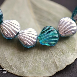 May include: A strand of teal and silver glass beads in the shape of seashells. The beads are arranged in a circle and are lying on a green leaf.