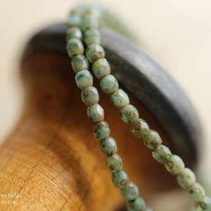 May include: Close-up of two strands of light green, faceted beads with brown and yellow accents. The beads are strung together, creating a delicate necklace or bracelet. The background is a blurred wooden surface. The text "... arte bella ... 10329" is visible.