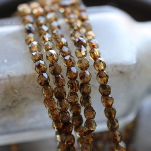May include: A strand of brown glass beads with a faceted, geometric shape. The beads have a translucent, amber-like appearance with flecks of gold and brown.