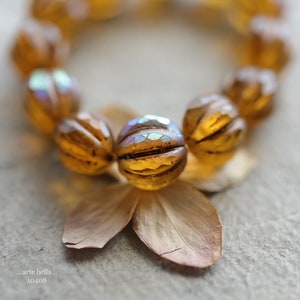 May include: A close-up of a brown and gold beaded necklace with iridescent accents. The beads are round and have a textured surface. The necklace is lying on a white surface.