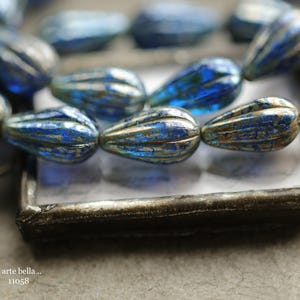 May include: Close-up of a strand of blue teardrop-shaped beads with a metallic finish. The beads have a ribbed texture and are displayed in a rectangular metal tray. The text ".. arte bella .. 11058" is visible.