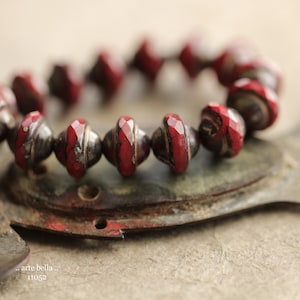 May include: Close-up of a beaded bracelet featuring red and silver-toned beads. The beads have a faceted design with a distressed finish. The bracelet is resting on a textured, metallic surface. Text in the image reads ".. arte bella.. 11052".