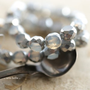 May include: A close-up of a silver and white glass bead necklace. The beads are faceted and have a metallic sheen. The necklace is resting on a silver spoon.