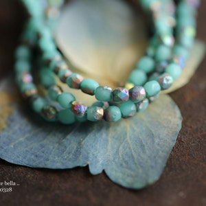 May include: A close-up of a multi-strand necklace made of turquoise blue glass beads. The beads have a faceted, iridescent finish, creating a shimmering effect. The necklace is resting on a light blue, textured surface.