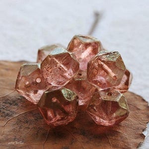 Op de afbeelding: Een verzameling gefacetteerde, roze glazen kralen. De kralen hebben een geometrische vorm met meerdere zijden en gaten om te rijgen. Ze liggen op een bruin blad, met de tekst "arte bella" linksonder.