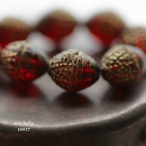 May include: Close-up of a strand of red and gold beads. The beads have a textured surface and a metallic gold finish. The image also includes the text ".. arte bella .. 10017".