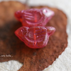 May include: Two pink glass bird-shaped beads with detailed carvings on a brown background.