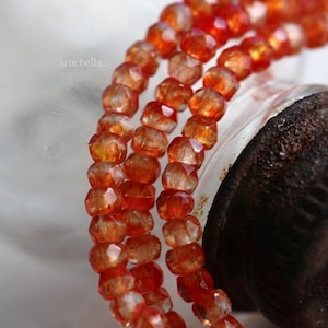 May include: Close-up of three strands of faceted orange gemstone beads. The beads are strung together, creating a layered effect. The beads are a warm orange color, with a faceted surface that catches the light. The text ".arte bella." is in the upper left corner.