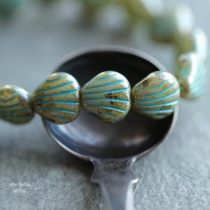 May include: A close-up of a strand of green and blue glass beads shaped like seashells. The beads are arranged in a circle and are resting on a silver spoon.