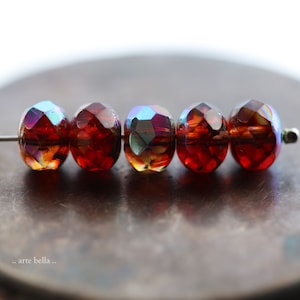 May include: Five iridescent red glass beads with a faceted design. The beads are strung on a wire and are arranged in a row.