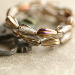 May include: Close-up of a strand of teardrop-shaped beads. The beads are clear with gold and purple iridescent stripes. The beads are arranged in a circular pattern. The text "arte bella.. 11060" is visible in the bottom left corner.