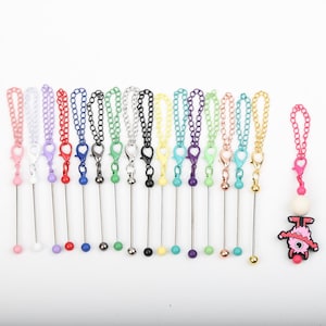 May include: A collection of colorful keychains with various chain colors and clasp colors. Each keychain has a metal rod with a decorative bead at the bottom. One keychain features a cartoon monster charm.