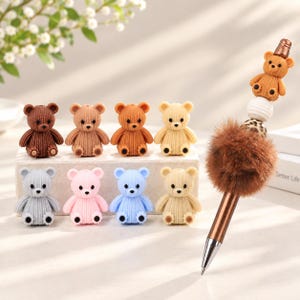 Könnte beinhalten: Eine Sammlung von acht kleinen, strukturierten bärenförmigen Perlen in verschiedenen Farben wie Braun, Rosa, Blau und Creme, sowie ein Stift mit einem braunen Kunstfell-Pompon, einer Leopardenmuster-Perle, einer weißen Perle und einer Bärenperle.