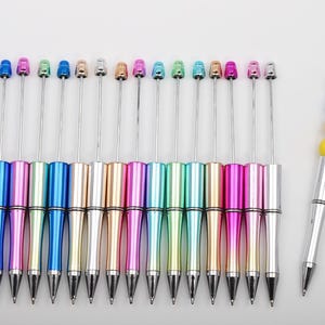May include: A set of 17 colorful pens with silver metal barrels and colorful tops. The pens have a variety of colors, including pink, blue, green, and silver. The pens are arranged in a row on a white surface.