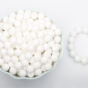 Pode incluir: Uma tigela azul clara cheia de contas brancas e redondas, juntamente com uma pulseira de contas brancas. As contas têm um acabamento fosco. A pulseira é feita de contas semelhantes. O fundo é branco liso.