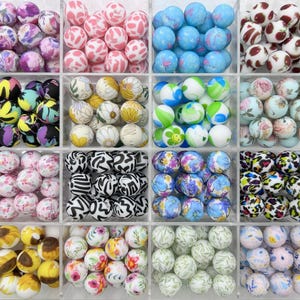 Pode incluir: Uma coleção de 12 contas de silicone com padrões diferentes em um recipiente de plástico transparente. As contas são organizadas em uma grade e apresentam vários designs, incluindo padrões florais, de estampa animal e abstratos.
