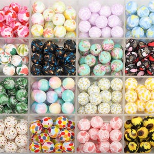 May include: A clear plastic organiser filled with various colourful beads. The beads are round and come in a variety of colours and patterns, including floral, marbled, and speckled designs. The beads are arranged in separate compartments.