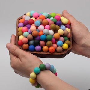 Pode incluir: Uma tigela de madeira cheia de uma variedade de contas redondas coloridas em vários tons. Uma pulseira feita de contas semelhantes é usada no pulso. As contas são em tons de rosa, azul, verde, laranja, amarelo e roxo.