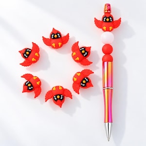 Könnte beinhalten: Ein Set aus sechs kleinen roten vogelformigen Silikonperlen und einem Stift. Der Stift hat einen metallisch irisierenden Schaft, einen roten Vogelaufsatz und zwei runde Perlen in Weiß und Rot. Jeder Vogel hat schwarze Augen und einen gelben Schnabel.
