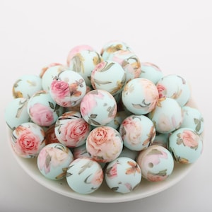 May include: A collection of round beads with a floral design. The beads are light blue with pink, peach, and green floral patterns. The beads are piled on a white plate.