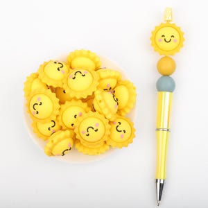 May include: A cheerful yellow pen with a smiling sun-shaped topper and a collection of matching sun-shaped embellishments. The pen has a silver tip and is decorated with yellow, orange, and blue beads. The embellishments are on a white plate.