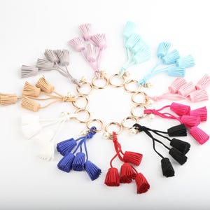 May include: A selection of tassel keyrings in a variety of colours, including pink, blue, black, red, and white. Each keyring has a gold-coloured ring and a small pearl detail. The tassels are made of a soft material. The keyrings are arranged in a circular pattern.
