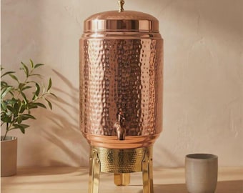 May include: A tall, cylindrical copper beverage dispenser with a hammered texture, a matching lid with a small knob, and a metal spigot. The dispenser rests on a gold-toned metal stand with three legs and includes a small, plain ceramic cup.