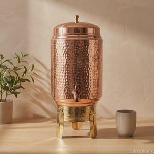 May include: A tall, cylindrical copper beverage dispenser with a hammered texture, featuring a matching lid with a small knob, a metal spigot, and a gold-toned metal stand with three legs. The dispenser is positioned next to a small ceramic cup.