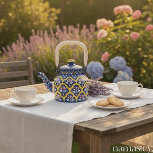May include: A blue and yellow patterned teapot with a cream-colored handle sits on a wooden table set for tea. Two white teacups and saucers, along with a plate of cookies, complete the scene. The background features a garden with flowers.