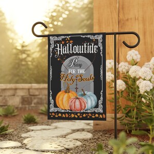May include: A black rectangular garden flag featuring a white ornate border and the text Hallowtide Pray for the Holy Souls. The design includes a gray tombstone with a rosary draped over it, flanked by three pumpkins in orange, red, and blue.