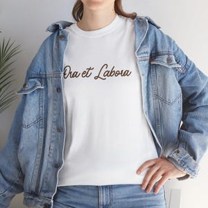 May include: White t-shirt with the brown text "Ora et Labora". The shirt is worn with a light blue denim jacket and blue jeans. The t-shirt is made of a soft, comfortable material.