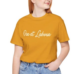 May include: Mustard yellow t-shirt with the cursive text "Ora et Labora" in white. The shirt has short sleeves and a crew neck. The model is wearing blue jeans.