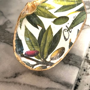 May include: A white seashell dish with a gold rim and a green and white olive branch design. The dish has the word "Olive" written on it in black cursive.