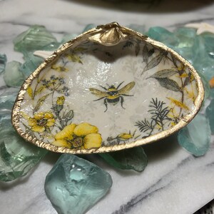 May include: A decorative seashell dish with a gold-painted rim. The interior features a floral and bee design with yellow and gray accents. The dish is resting on a bed of blue-green glass pebbles.