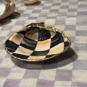 May include: A small, white seashell with a black and gold checkerboard pattern painted on the inside. The shell is sitting on a white and purple checkered surface.