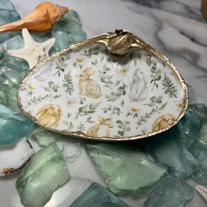 May include: A white seashell dish with a gold rim and a floral pattern featuring bunnies and leaves. The dish is sitting on a surface with green and blue glass pieces.