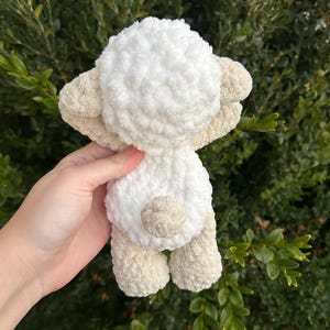 Puede incluir: Un juguete de oveja de peluche hecho a mano en crema y beige. La oveja tiene un cuerpo blanco esponjoso, orejas, patas y una peque&ntilde;a cola beige. El juguete se sostiene contra un fondo de follaje verde.