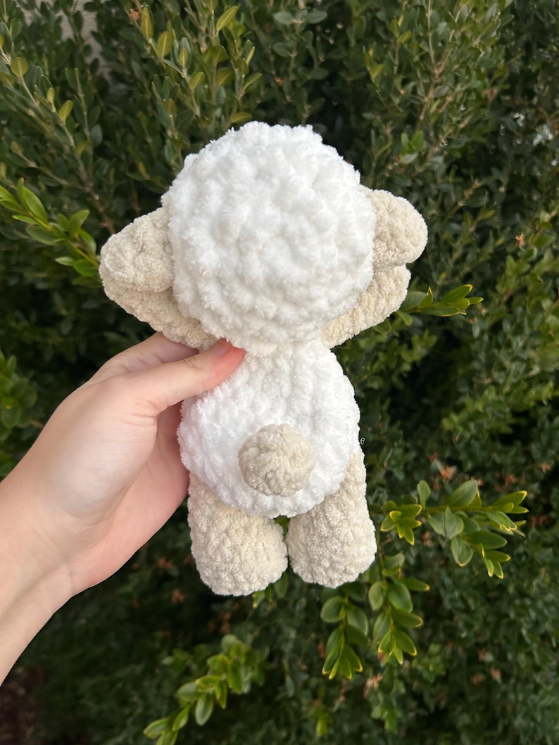 Puede incluir: Un juguete de oveja de peluche hecho a mano en crema y beige. La oveja tiene un cuerpo blanco esponjoso, orejas, patas y una peque&ntilde;a cola beige. El juguete se sostiene contra un fondo de follaje verde.