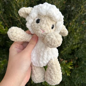 Puede incluir: Un peluche de cordero hecho a mano con ganchillo. El cordero tiene una cabeza y un cuerpo blancos y esponjosos, con patas y brazos beige. Tiene ojos negros y una boca cosida rosa. El juguete se sostiene sobre un fondo de follaje verde.