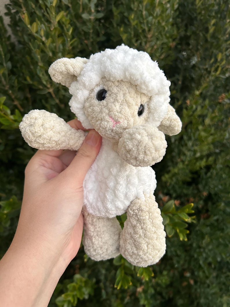 Puede incluir: Un peluche de cordero hecho a mano con ganchillo. El cordero tiene una cabeza y un cuerpo blancos y esponjosos, con patas y brazos beige. Tiene ojos negros y una boca cosida rosa. El juguete se sostiene sobre un fondo de follaje verde.
