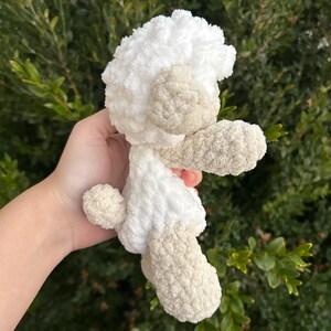 Puede incluir: Un peluche de cordero de ganchillo hecho a mano en blanco y beige. El cordero tiene una cabeza y un cuerpo blancos y esponjosos, con detalles beige en las orejas, las patas y la cola. El peluche es suave y tierno.