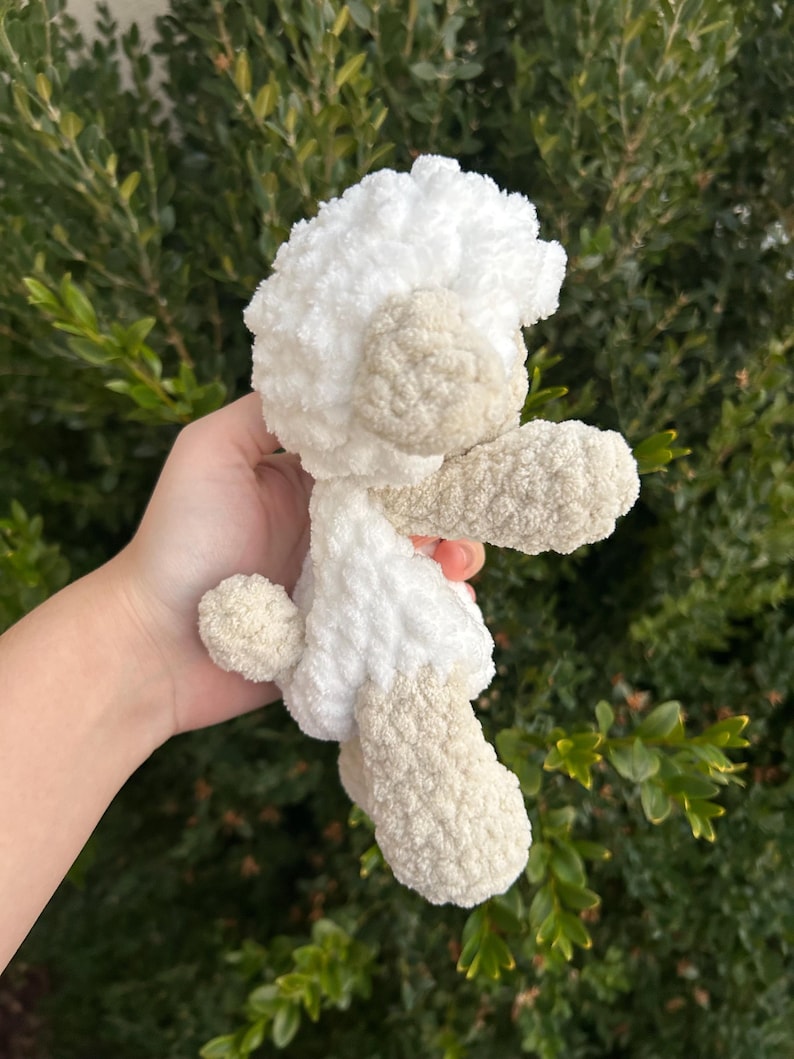 Puede incluir: Un peluche de cordero de ganchillo hecho a mano en blanco y beige. El cordero tiene una cabeza y un cuerpo blancos y esponjosos, con detalles beige en las orejas, las patas y la cola. El peluche es suave y tierno.