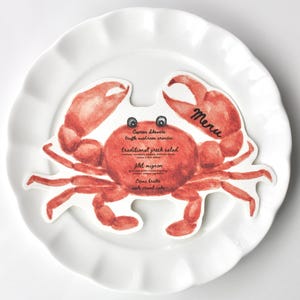 May include: A red crab-shaped menu card with the word "Menu" printed in black. The menu lists: Caprese skewers, truffle mushroom panini, traditional Greek salad, filet mignon, creme brulee, apple strudel cake.