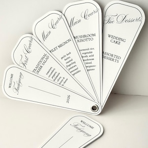 May include: A set of white fan-shaped menu cards with black text and a metal grommet. The cards list courses including First Course, Main Course, and The Desserts, with specific food items like Greek Salad, Filet Mignon, and Mushroom Risotto.