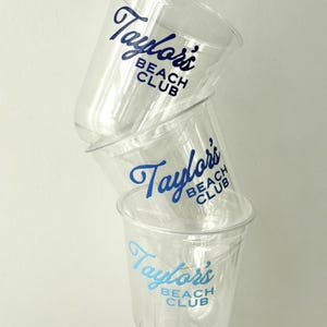 May include: A stack of three clear plastic cups with the text "Taylor's Beach Club" printed in blue. The cups are slightly transparent, and the text is in a cursive font. The cups are stacked on top of each other.