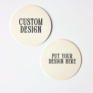 May include: Two circular, off-white coasters with black text. One says "CUSTOM DESIGN," the other "PUT YOUR DESIGN HERE." The coasters are made of a textured material and are on a white background.